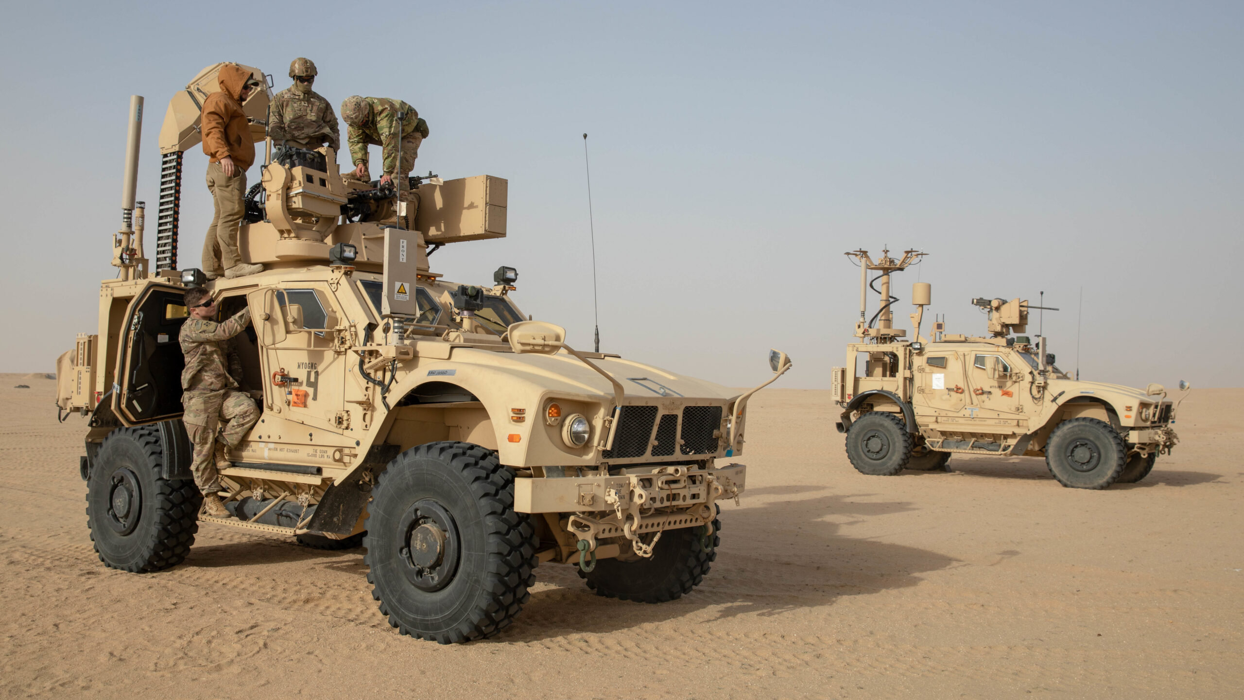 Soldiers work atop an M-LIDS-equipped armored vehicle mounted with a counter-drone sensor and weapon turret in a desert training exercise, with a second equipped vehicle in the background
